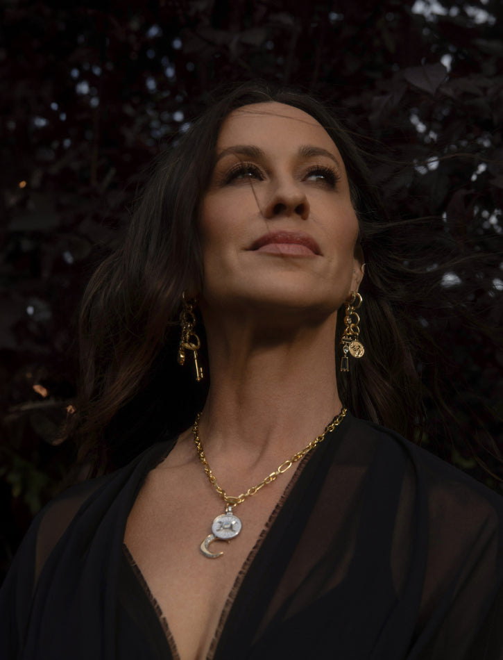 Product image of A woman with long dark hair wears gold jewelry, including dangling earrings and a necklace, and gazes upward. She is dressed in a black top. The background is dark, possibly with foliage.