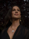 A woman with long dark hair wears gold jewelry, including dangling earrings and a necklace, and gazes upward. She is dressed in a black top. The background is dark, possibly with foliage.