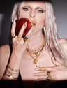 A person with light hair holds a red apple and wears multiple gold necklaces, bracelets, rings, and a black top. The background is neutral, and the lighting casts shadows on the face and the apple.