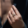 A close-up of a hand adorned with the Infinity Snake Ring by Awe Inspired, featuring white topaz accents and shaped like two intertwined leaves, with a dark fabric visible in the background.