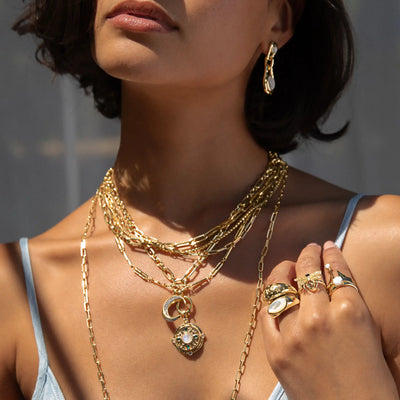 A person adorned with multiple gold necklaces, rings, and earrings, holding an Awe Inspired Manifestation Locket.