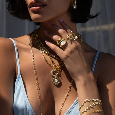 Close-up of a woman showcasing layered gold necklaces, including the Manifestation Locket by Awe Inspired, along with elegant rings and a bracelet, all adorned with intricate designs and moonstones. She is dressed in a light blue V-neck top, gently touching her neck and jewelry.