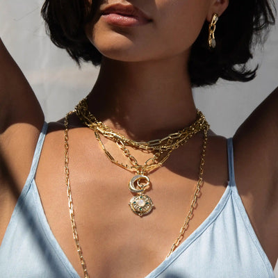 Close-up of a woman wearing gold necklaces and earrings, with a blue sleeveless top. The necklaces feature intricate pendants, including the Manifestation Locket by Awe Inspired with a moonstone centerpiece. The lighting casts shadows on her skin, hinting at a secret desire hidden within.