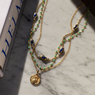 A Lapis Turquoise Choker Necklace layered with standard Aphrodite pendant and Colored Enamel necklace by Awe Inspired on a marble table next to a book.