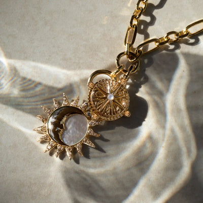 The True North Charm Necklace by Awe Inspired features a gold necklace with a sun-shaped pendant and an ornate circular Sun & Moon amulet, both hanging from a chain, casting shadows on a light grey surface.