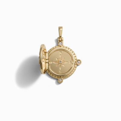 The True North Charm Necklace by Awe Inspired is a gold locket with a compass design on its front cover, partially open to reveal an empty interior. The locket features small gemstones, Mother of Pearl accents, and a loop for a chain.