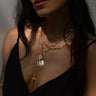 A woman wearing layered gold necklaces with various pendants, including an Awe Inspired Compass Locket, a sun, and a sword, partially visible in a dark sleeveless top.