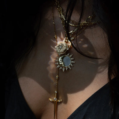 Close-up of a person wearing the Awe Inspired True North Charm Necklace, featuring a layered gold design with a sun-like pendant and drop charm over a dark top, elegantly showcasing an exquisite Mother of Pearl inlay.