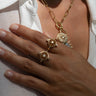 A hand with light skin tone adorned with two Awe Inspired Compass Rose Rings featuring intricate compass designs and a matching necklace with a mother of pearl center, all worn over a white button-down shirt.