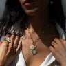 A woman accessorized with Awe Inspired's Compass Locket featuring celestial pendants, along with several gold rings, gently holding her collar. Her dark hair drapes over a white garment.