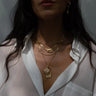 A person wearing a sheer white blouse showcases the Inner Compass Necklace Set by Awe Inspired along with a gold earring, symbolizing prosperity, with dark hair cascading over the shoulders.