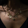 Close-up of a person wearing the Awe Inspired Compass Locket, a gold chain necklace adorned with charms like a sun, a star, a crescent moon, and other intricate pendants such as a locket and a gemstone-studded compass rose, against a dark background.