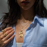 A woman wearing the Awe Inspired "We Are The Future Necklace Set," including a Kamala pendant, along with a blue and white striped shirt and several rings, is partially visible from the shoulders up.