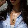 Close-up of a person wearing the Reach New Heights Necklace Set by Awe Inspired, featuring layered gold necklaces that include a Kamala Harris pendant and a small charm, over a blue and white striped button-up shirt. The person's face is partially visible.