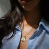 A person wearing a blue striped shirt showcases the "Votes for Women Kamala Necklace Set" by Awe Inspired, featuring multiple gold pendants. The set includes a detailed relief pendant, a padlock-shaped charm, and a key. Notably, it includes a Kamala Harris pendant celebrating the women's suffrage movement.