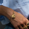 Close-up of a hand adorned with the Vote for Kamala Bracelet Set from Awe Inspired, featuring a coin charm and several gold rings. The person is dressed in a blue and white striped shirt paired with denim jeans.