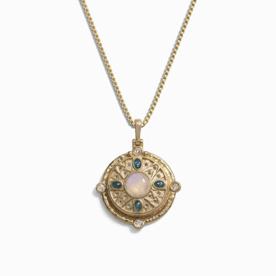 The Manifestation Locket Necklace by Awe Inspired showcases a gold pendant with an intricate circular design, highlighting a central moonstone encircled by smaller blue and clear stones, all elegantly attached to a gold chain.