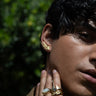 Close-up of a person wearing a gold wing-shaped earring and multiple rings on their fingers, one being the striking Opal Crown Ring from Awe Inspired, featuring black mother of pearl. A hand gently touches the face, framed by lush green foliage in the background.