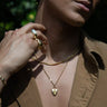 A person wearing layered Miami Cuban Chain necklaces by Awe Inspired, including a layering chain and one with a heart-shaped pendant, along with several rings, while holding the collar of their open brown shirt.