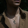 A person wearing an open-collared brown shirt and the Sea & Sky Charm Necklace from Awe Inspired, featuring a gold chain and a pendant shaped like an abstract flower made of black mother of pearl. The background is dark and out of focus.