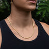 A person wearing a black tank top and an Awe Inspired Miami Cuban Chain necklace. The background is slightly blurred with green foliage.