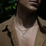 A person wearing layered gold and silver necklaces, including the Awe Inspired Twisted Moon Necklace adorned with white topaz, paired with a brown button-up shirt against a dark, leafy background.