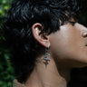 Close-up profile of a person with dark, wavy hair wearing an Awe Inspired Snake Eyes Charm Earring in their left ear. The background, blurred greenery, helps refresh your outlook.