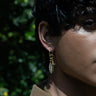 A person wears a Pen & Sword Charm Earring from Awe Inspired, showcasing a Mother of Pearl Feather and sword charm, with a blurred green background.