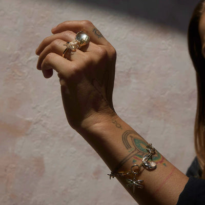 A hand adorned with various tattoos, wearing a large, clear ring and an Awe Inspired Earth Magic Charm Bracelet featuring a lucky dragonfly, is raised and partially in shadow.