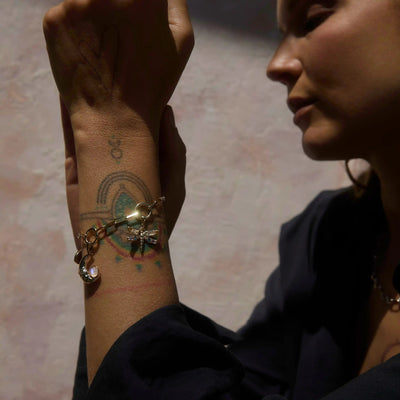 Close-up image of a person highlighting their wrist adorned with an Earth Magic Charm Bracelet from Awe Inspired. The individual has tattoos on their wrist and is dressed in dark clothing, set against a light-colored wall.