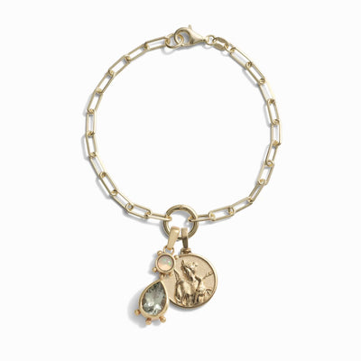 Awe Inspired's Wild Goddess Charm Bracelet features a gold chain with three dangling charms: a bezel set gemstone, a coin engraved with the Goddess of the Wilds, and a small round stone.