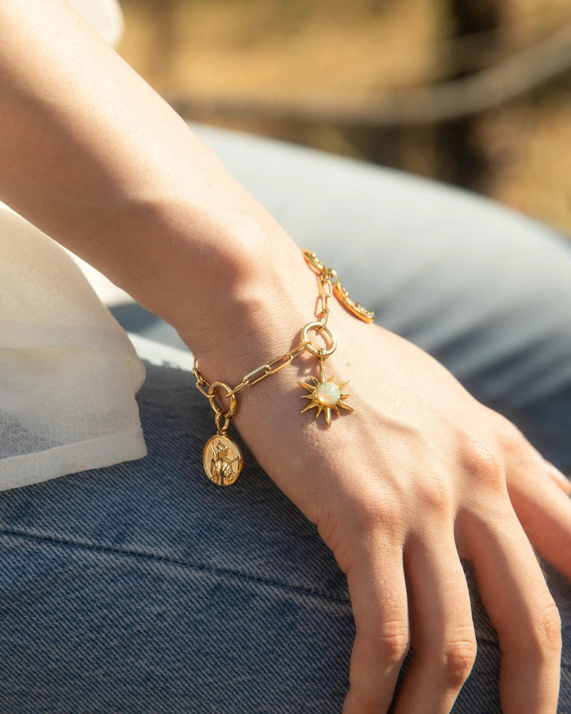 A close-up of a person's wrist adorned with the "Cosmic Euphoria Charm Bracelet" by Awe Inspired, featuring a gold bracelet with a sun-shaped charm and other small pendants, including an opal sun, resting on their blue jeans.