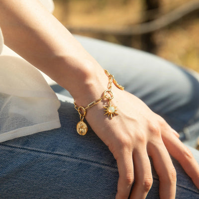 A close-up of a person's wrist adorned with the "Cosmic Euphoria Charm Bracelet" by Awe Inspired, featuring a gold bracelet with a sun-shaped charm and other small pendants, including an opal sun, resting on their blue jeans.