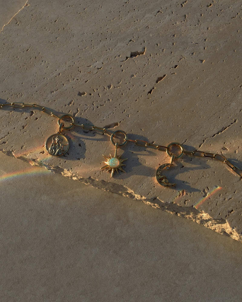 The Cosmic Euphoria Charm Bracelet by Awe Inspired, featuring a gold chain with three charms—a circular coin, a starburst adorned with a white gem, and a crescent moon—is elegantly displayed on textured stone. Subtle rainbow reflections cast by the light nearby enhance the cosmic euphoria feel.