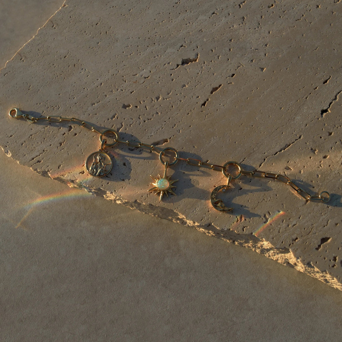 The Cosmic Euphoria Charm Bracelet by Awe Inspired, featuring a gold chain with three charms—a circular coin, a starburst adorned with a white gem, and a crescent moon—is elegantly displayed on textured stone. Subtle rainbow reflections cast by the light nearby enhance the cosmic euphoria feel.
