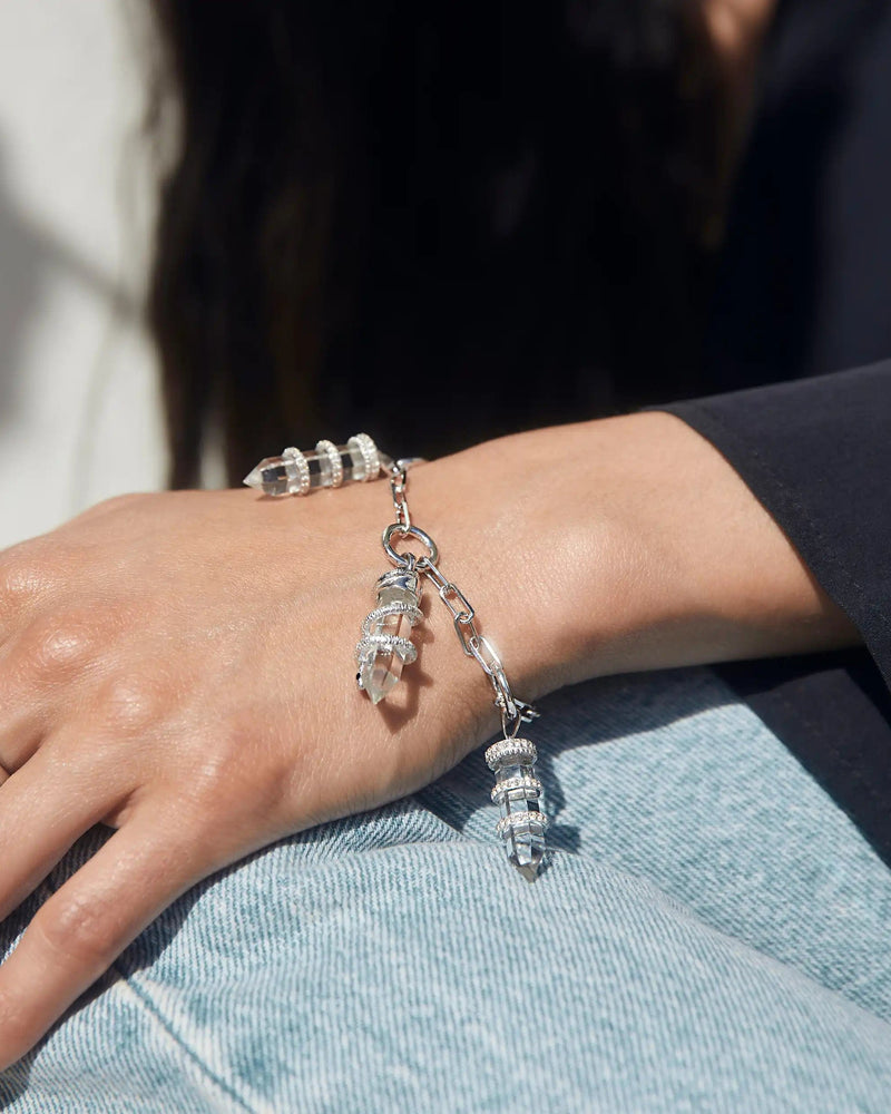 Close-up of a person wearing the Guardian's Crystal Charm Bracelet by Awe Inspired, featuring clear, cylindrical beads reminiscent of quartz crystals, alongside several silver rings on their fingers, resting their hand on their leg in blue jeans.
