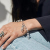 Close-up of a person wearing the Guardian's Crystal Charm Bracelet by Awe Inspired, featuring clear, cylindrical beads reminiscent of quartz crystals, alongside several silver rings on their fingers, resting their hand on their leg in blue jeans.