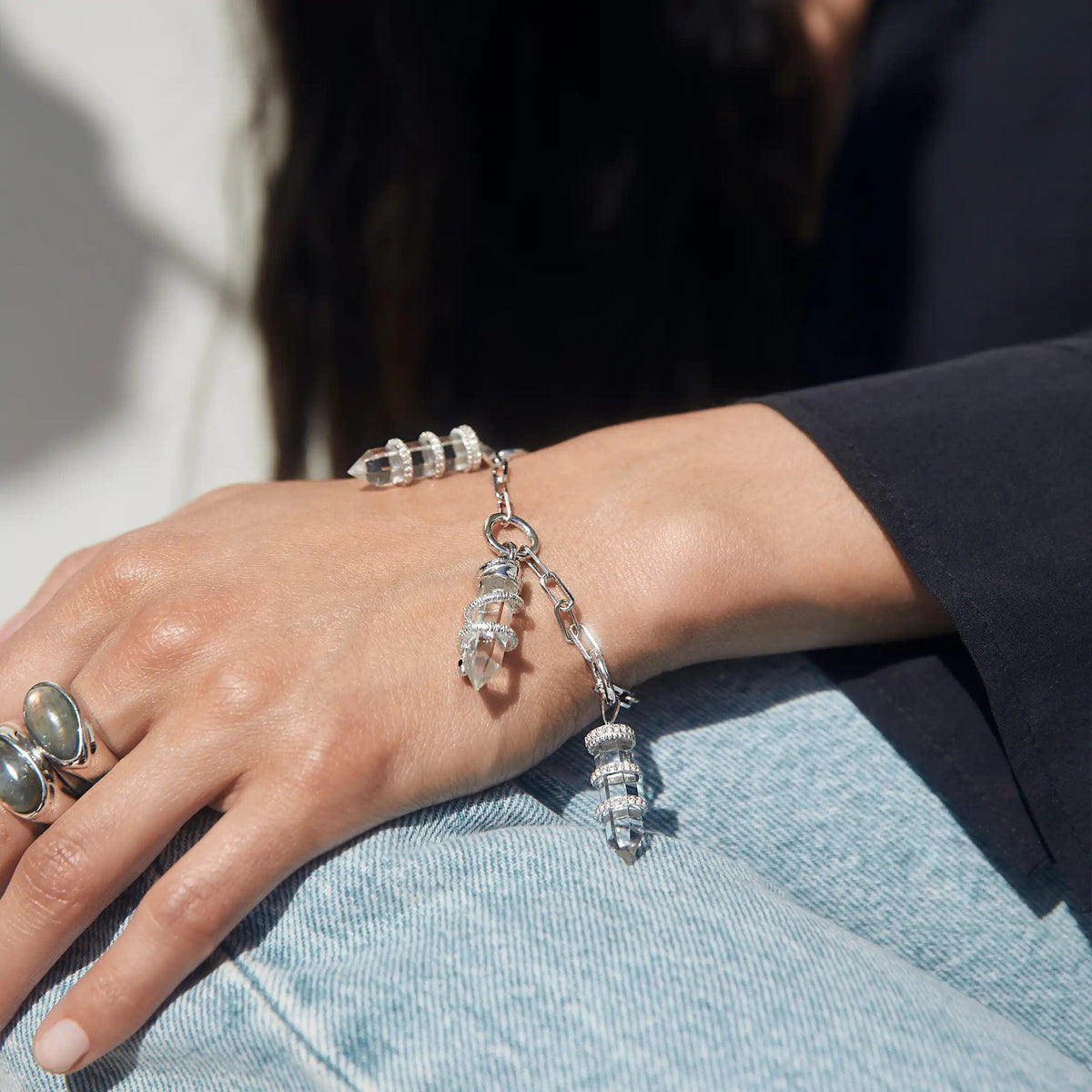 Close-up of a person wearing the Guardian's Crystal Charm Bracelet by Awe Inspired, featuring clear, cylindrical beads reminiscent of quartz crystals, alongside several silver rings on their fingers, resting their hand on their leg in blue jeans.