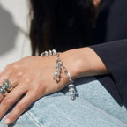 Close-up of a person wearing the Guardian's Crystal Charm Bracelet by Awe Inspired, featuring clear, cylindrical beads reminiscent of quartz crystals, alongside several silver rings on their fingers, resting their hand on their leg in blue jeans.