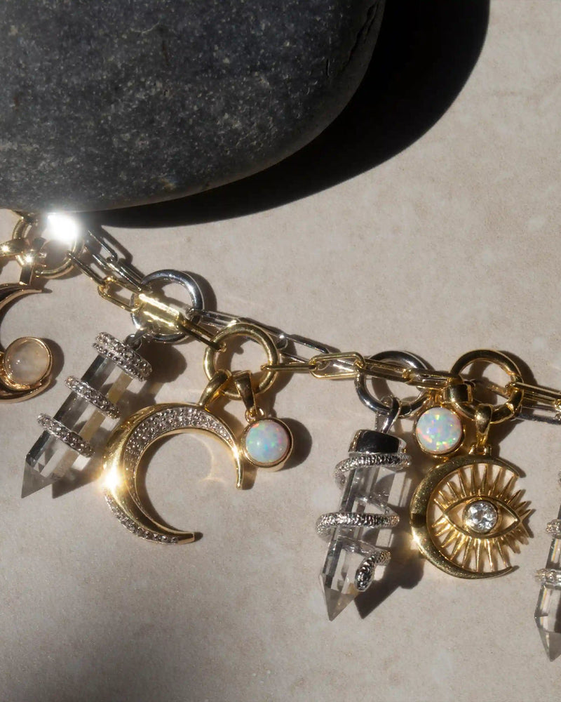 A Guardian's Crystal Charm Bracelet by Awe Inspired, adorned with gold moon and sun charms, crystal pendants, and opals, rests on a light surface next to a quartz crystal.