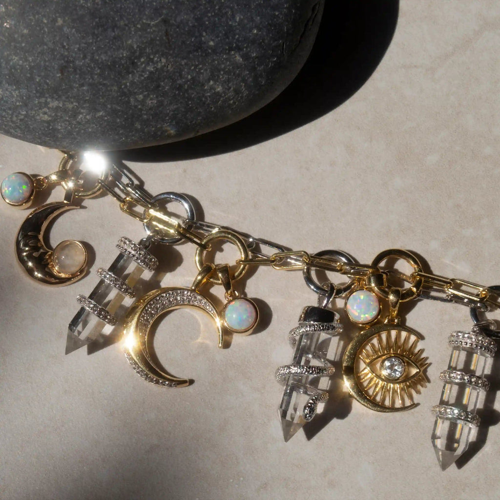 A Guardian's Crystal Charm Bracelet by Awe Inspired, adorned with gold moon and sun charms, crystal pendants, and opals, rests on a light surface next to a quartz crystal.