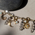 A Guardian's Crystal Charm Bracelet by Awe Inspired, adorned with gold moon and sun charms, crystal pendants, and opals, rests on a light surface next to a quartz crystal.