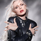 Person with long platinum blonde hair and dramatic makeup poses with hands crossed in front, wearing a black leather outfit adorned with multiple rings and jewelry, including a Guardian's Crystal Charm Bracelet by Awe Inspired featuring white sapphires, against a grey background.
