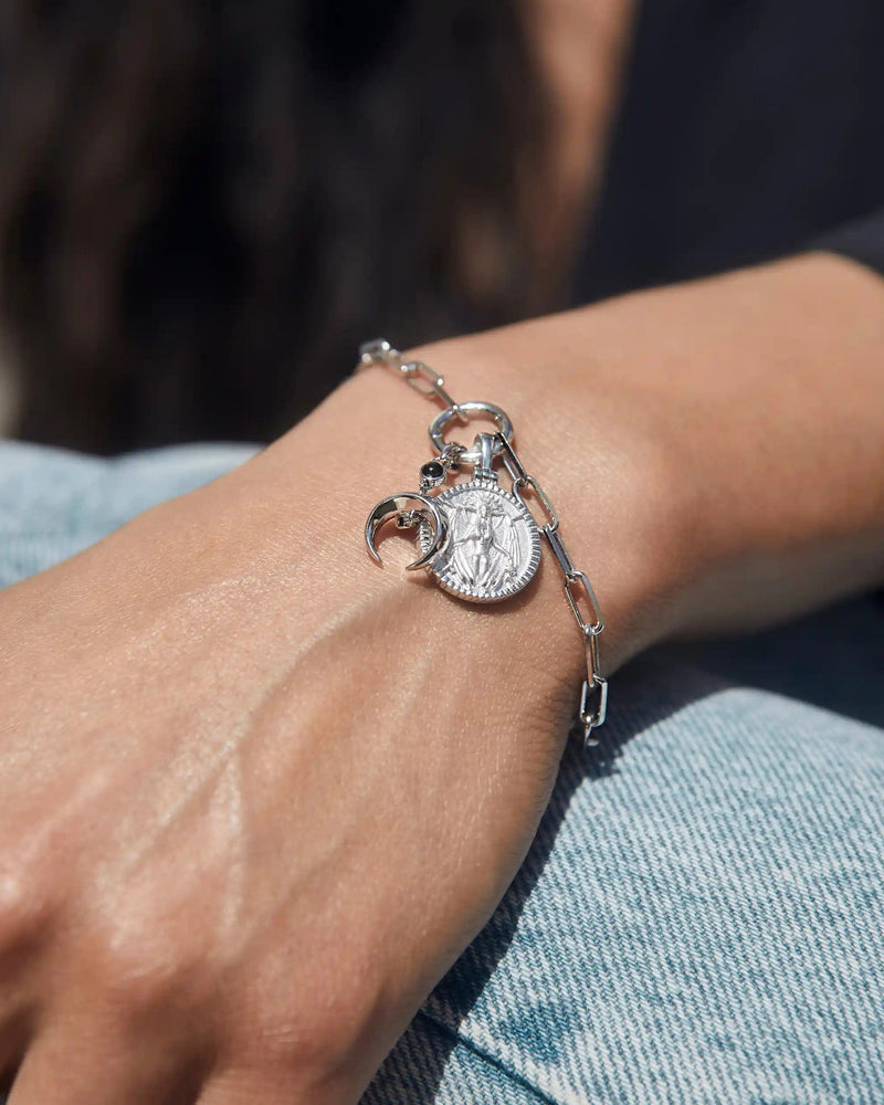 A hand wearing the Awe Inspired Inverted Moon Charm Bracelet, featuring a silver chain adorned with a coin pendant and two black moon charms, rests on denim jeans.