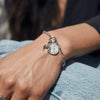 A hand wearing the Awe Inspired Inverted Moon Charm Bracelet, featuring a silver chain adorned with a coin pendant and two black moon charms, rests on denim jeans.