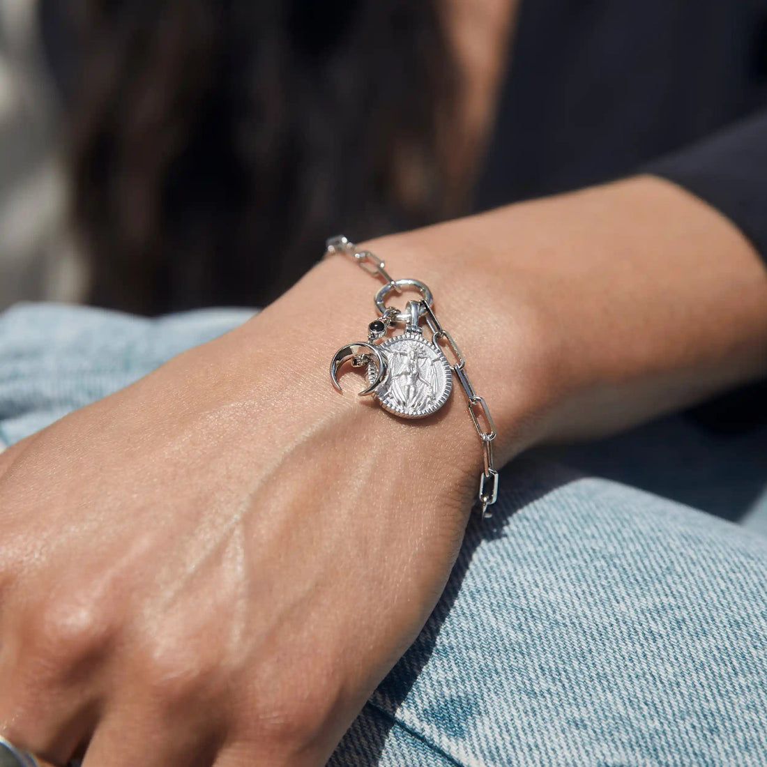 A hand wearing the Awe Inspired Inverted Moon Charm Bracelet, featuring a silver chain adorned with a coin pendant and two black moon charms, rests on denim jeans.