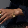 A close-up of a person's wrist wearing the Inverted Moon Charm Bracelet from Awe Inspired, showcasing its multiple pendants including a striking black moon. The person is also adorned in gold rings and dressed in a dark long-sleeve shirt.
