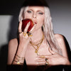 A person with long hair bites into an apple while wearing the Inverted Moon Charm Bracelet by Awe Inspired, alongside multiple gold necklaces, rings, and other bracelets. A pendant hangs around their neck. The background is plain, and part of their attire is visible.