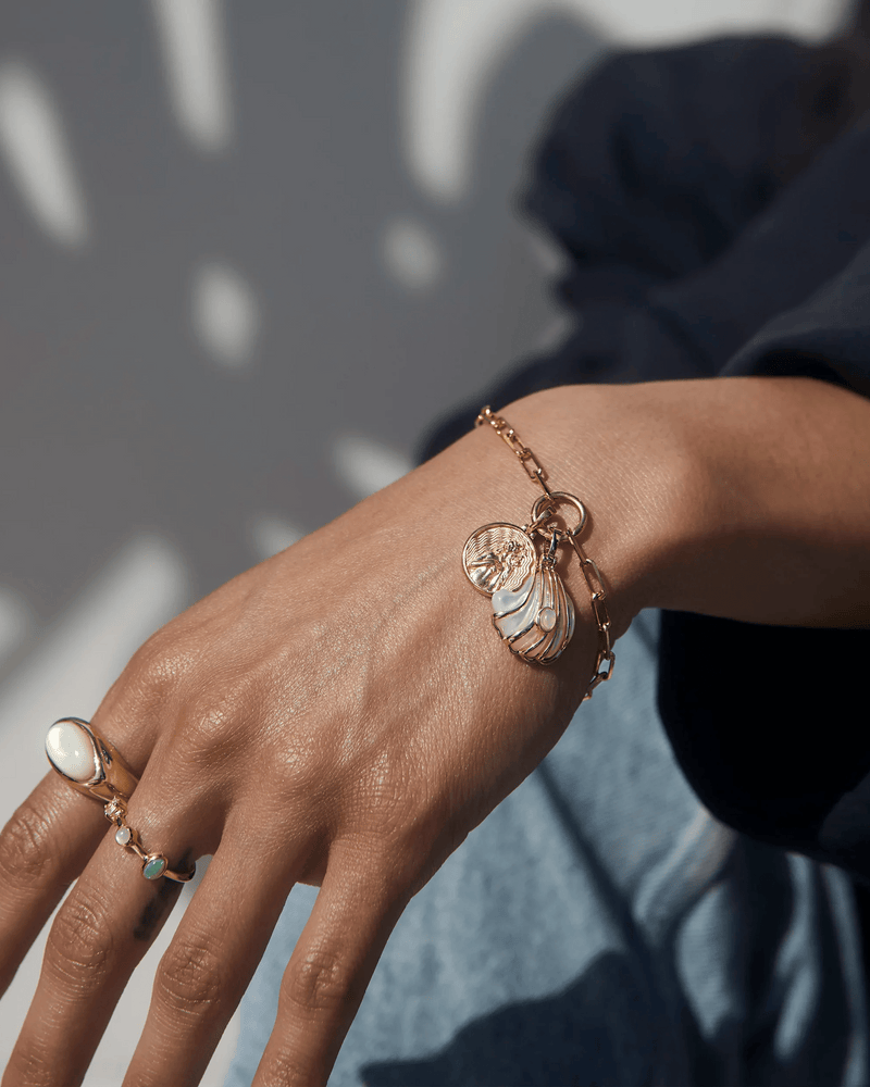 A close-up of a person's hand adorned with the Ocean Goddess Charm Bracelet from Awe Inspired and a ring featuring a large pearl. The person, dressed in a dark long-sleeve shirt, exudes beauty and elegance.