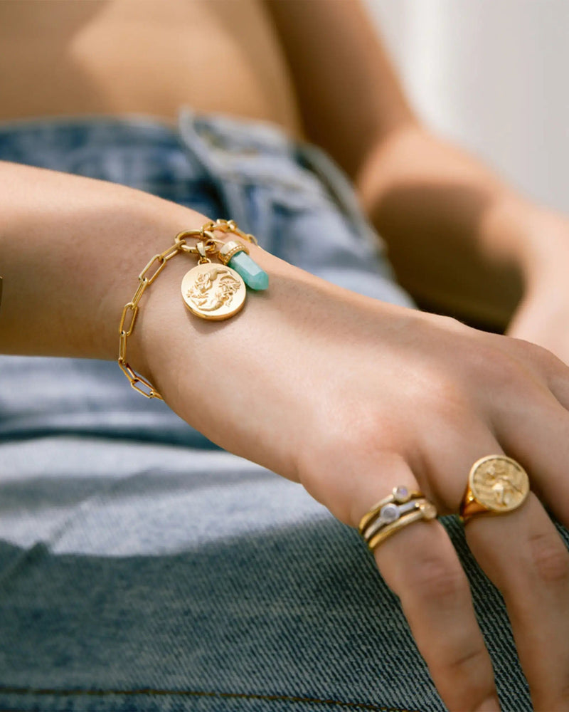 A person wearing a Single Charm Collector Bracelet by Awe Inspired adorned with gold charms and multiple rings on their fingers, resting their hand on their lap, dressed in denim.